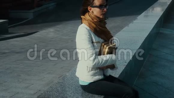 戴着眼镜手里拿着书的年轻黑发美女坐在街上视频的预览图