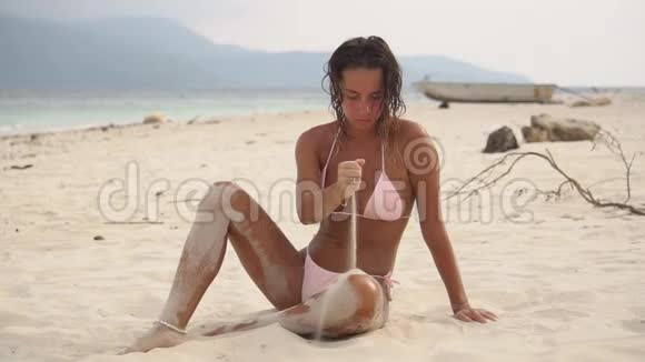 户外拍摄的美丽的年轻女子泳装她坐在沙滩上玩沙子视频的预览图