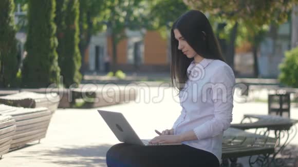 年轻女子坐在街上的长椅上一边用手提电脑键盘打字视频的预览图