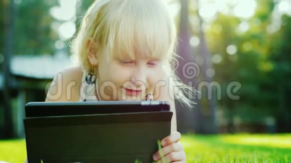 金发女孩6年在平板电脑上玩情绪反应躺在房子后院的草坪上视频的预览图