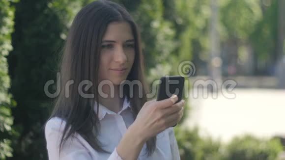 一个穿着白色衬衫的年轻女人的肖像下午在城市的一条街道上使用智能手机视频的预览图