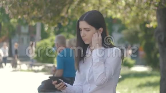 侧面观看白色衬衫年轻女子的肖像使用耳机和智能手机听音乐城市室外视频的预览图