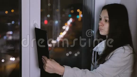 在黑暗的夜晚女人坐在窗台上在平板电脑上做自拍视频的预览图