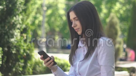 一个穿着白色衬衫的年轻女人的肖像下午在城市的一条街道上使用智能手机视频的预览图