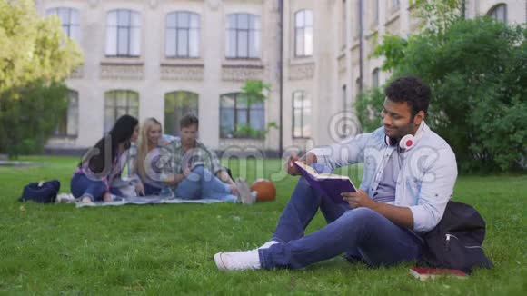 快乐的混血男学生坐在草地上读有趣的书视频的预览图