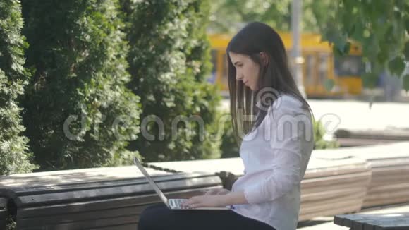 年轻女子坐在街上的长椅上一边用手提电脑键盘打字视频的预览图
