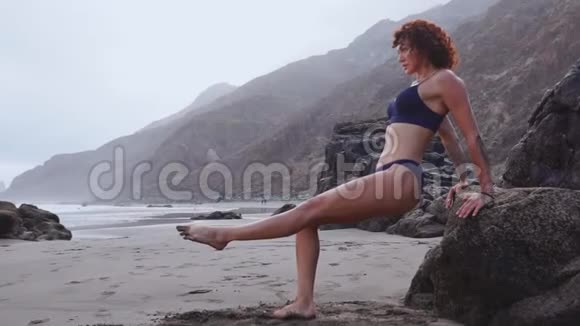 女人在靠近海洋的海滩上的岩石上做推UPS在海滩上做俯卧撑的年轻女性视频的预览图