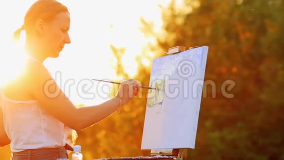 一个穿着白色t恤留着白发的女孩的肖像用油在夕阳下的画布上描绘了一幅风景视频的预览图