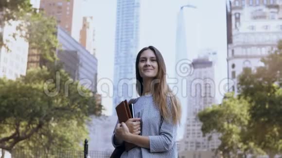 站在纽约金融区的年轻快乐女商人的肖像靠近摩天大楼拿着笔记本视频的预览图