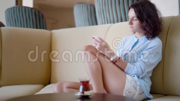 年轻漂亮的留着短波浪头的女人坐在沙发上用智能手机看照片视频的预览图