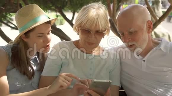 快乐的家庭享受假期老年人和他们的女儿坐在公园里浏览智能手机视频的预览图