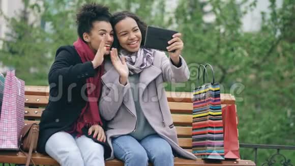 两个快乐的非裔美国妇女带着购物袋用平板电脑打视频电话年轻的朋友坐着视频的预览图