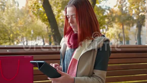在阳光明媚的天气里长着狐毛的女孩坐在长凳上使用平板电脑并在网上订购视频的预览图