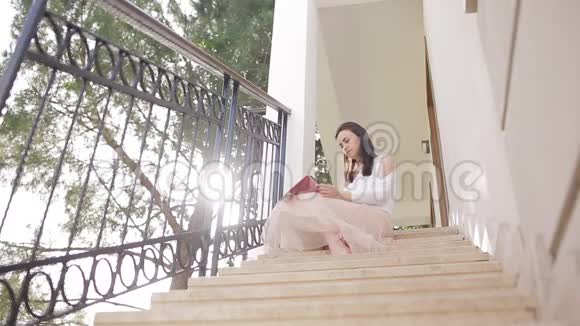 一位拿着数码平板电脑的年轻女子坐在靠近森林的房子前的楼梯上视频的预览图