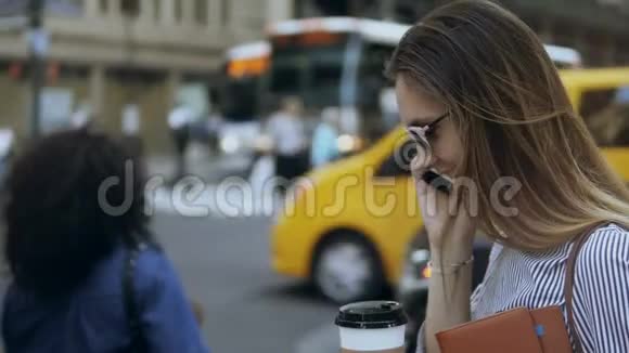 年轻漂亮的女商人拿着咖啡站在繁忙的街道上在路边的智能手机上交谈视频的预览图