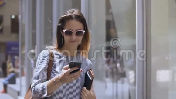 年轻漂亮的女商人戴着太阳镜拿着文件用智能手机走在街上慢动作视频的预览图