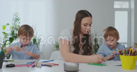 一个有两个孩子的家庭和一个坐在桌子旁的年轻母亲用彩色铅笔在纸上画画发展视频的预览图