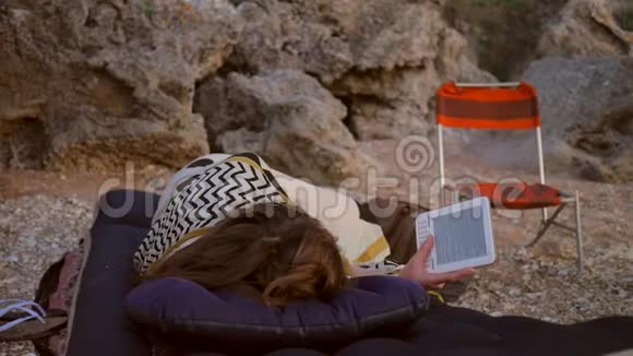 年轻的学生女孩躺在充气床垫露营和阅读电子书在海滩上高清视频的预览图