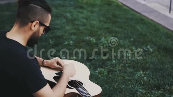 年轻的帅哥胡子拉碴坐在公园的草地上换着琴弦把吉他放在外面视频的预览图