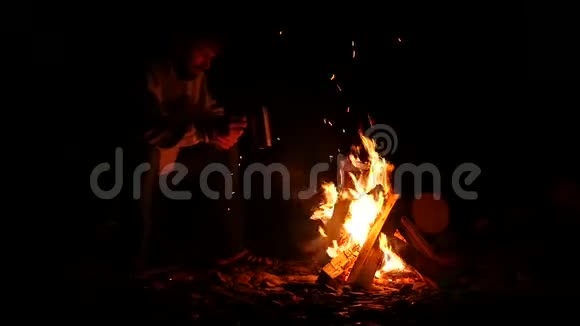大胡子的男人喝着大杯子里的茶在黑暗中在篝火的手机上拍张照片慢动作视频的预览图