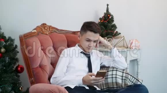 一个手里拿着智能手机的年轻人坐在靠近新年树的扶手椅上视频的预览图