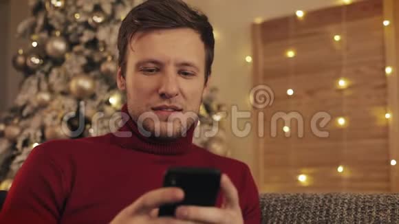 迷人的男人使用智能手机坐在沙发上发信息微笑在装饰好的公寓附近圣诞树视频的预览图