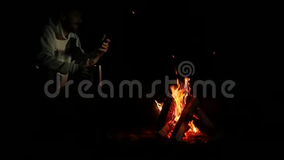 大胡子的男人喝着大杯子里的茶在黑暗中在篝火的手机上拍张照片慢动作视频的预览图
