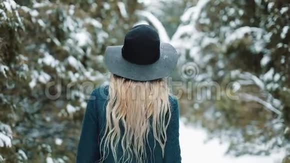从后面看着戴着绿色帽子和外套的女士在冬天的森林里走来走去女孩看着镜头摆姿势视频的预览图