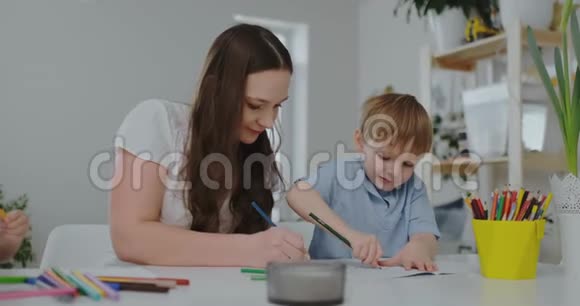 一个有两个孩子的家庭和一个坐在桌子旁的年轻母亲用彩色铅笔在纸上画画发展视频的预览图