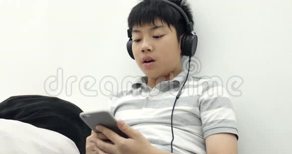 一个快乐的亚洲青少年男孩躺在清醒的智能手机和耳机和听音乐在家里的肖像视频的预览图