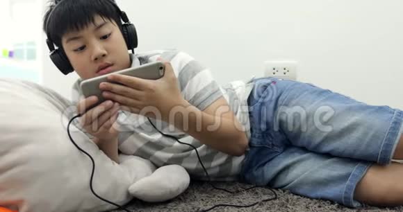 一个快乐的亚洲青少年男孩躺在清醒的智能手机和耳机和听音乐在家里的肖像视频的预览图