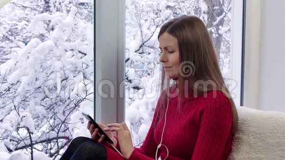 穿红裙子的漂亮女孩坐在窗台上用智能手机带耳机冬天在外面视频的预览图