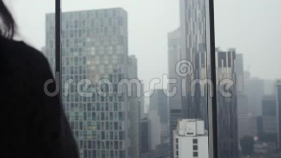 雨天的年轻女子站在窗边喝茶望着年的城市全景建筑视频的预览图