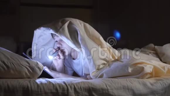 一个孩子晚上用手电筒在毯子下面读一本书热心男孩视频的预览图