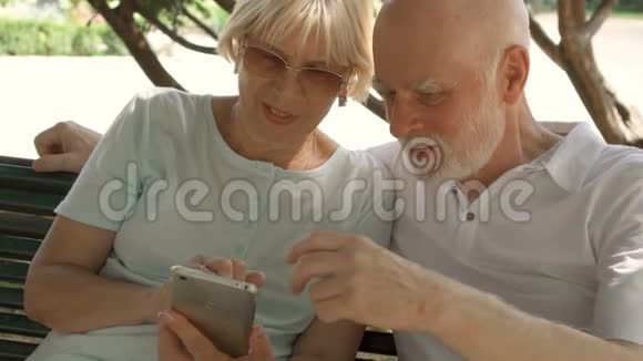 在欧洲度假的快乐家庭坐在公园长椅上的老年人在智能手机上浏览视频的预览图