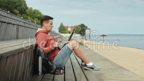 成熟的男人游客用笔记本电脑坐在沙滩上的木凳上视频的预览图