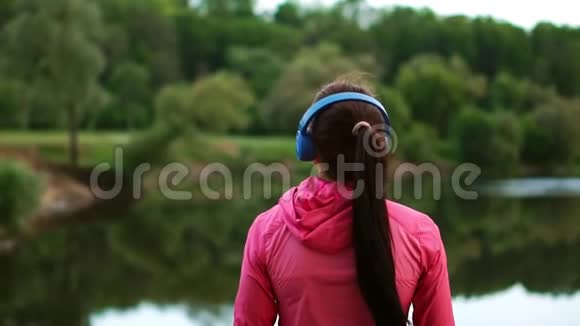 一个穿着粉红色夹克和蓝色耳机的女孩背着她站着一大早跑完后看着河视频的预览图