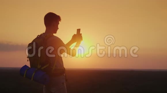 一个带背包的年轻人正在日落时拍摄一幅美丽的风景橙色天空背景的剪影视频的预览图