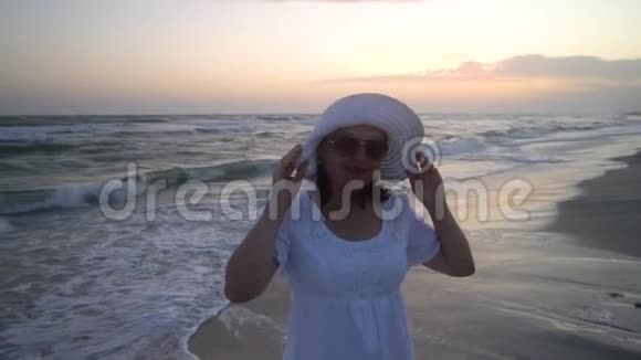 年轻漂亮的女人在日落时站在海边穿着白色连衣裙和白色帽子她看着相机微笑视频的预览图
