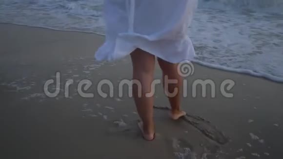 年轻漂亮的女人在日落时走在海边腿部特写穿着白色连衣裙和白色帽子视频的预览图