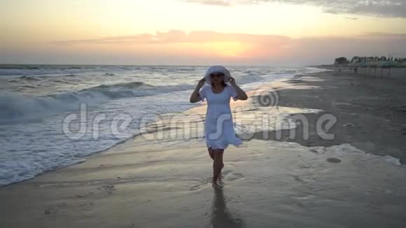 年轻漂亮的女人在日落时走在海边穿着白色连衣裙和白色帽子那个女人怀孕了她离开了视频的预览图