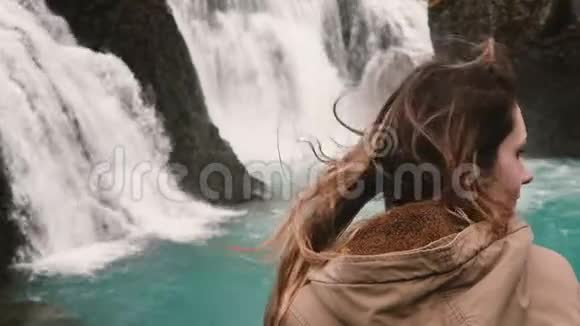 近距离观看年轻迷人的女人独自站立看着冰岛强大的瀑布风中的头发波浪视频的预览图