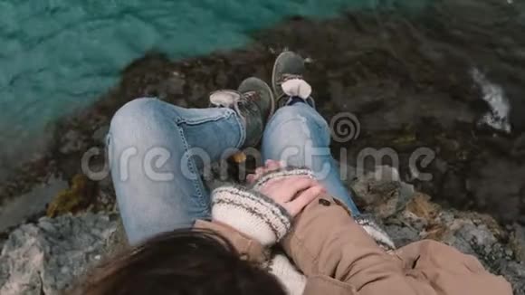 顶部的特写镜头年轻的黑发女人坐在岩石上看着蓝色的水头发在风中飘扬视频的预览图