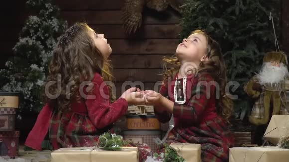 可爱的双胞胎穿着连衣裙坐在圣诞装饰中抬头看着飘落的雪慢动作视频的预览图