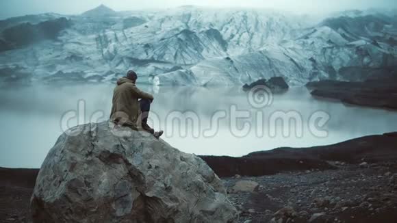 年轻的帅哥坐在冰泻湖的岩石上看着冰岛的冰川游客站起来走开视频的预览图