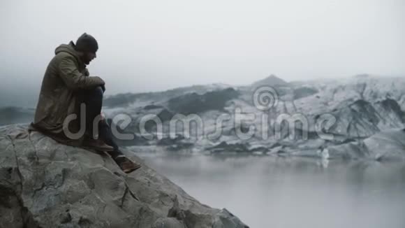 年轻英俊的男人坐在岩石上看冰川仅在冰岛探索冰泻湖的游客视频的预览图