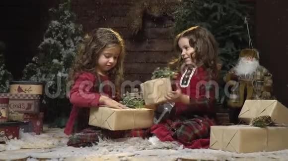 穿着红色衣服的可爱的姐妹们坐在房间里带着礼物房间里有圣诞装饰雪落在她们身上视频的预览图