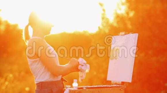 一个穿着白色t恤留着白发的女孩的肖像用油在夕阳下的画布上描绘了一幅风景视频的预览图