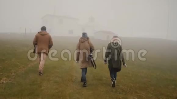 雾天三位年轻旅行者在山区山谷徒步旅行朋友们走过田野视频的预览图