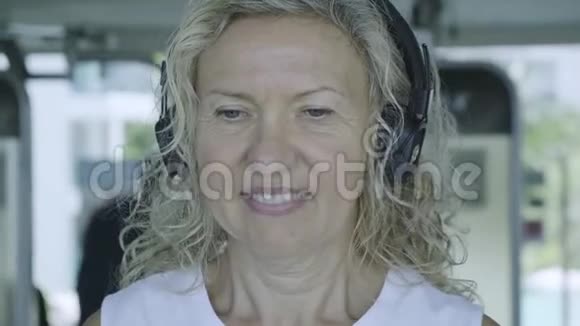 老年妇女在跑步机上健身房听音乐带耳机特写视频的预览图